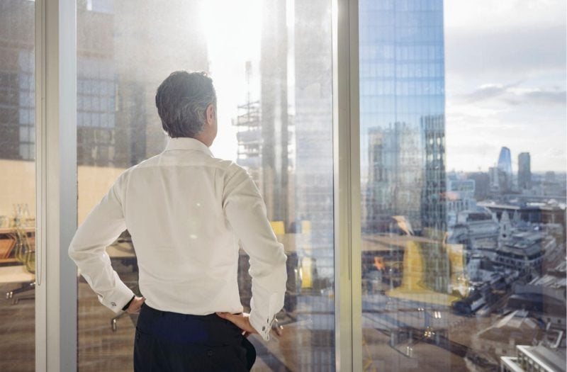 Business owner standing by office window looking out over city, reflecting during a moment of financial stress